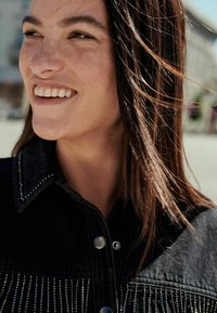 Sort denimskjorte med krave, sølvperle accenter og lodret frynsedetalje. Modellen har langt brunt hår og et naturligt smil.