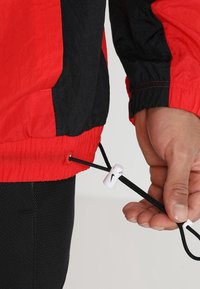 Giacca a vento rossa e nera con orlo elastico e cordino regolabile. Presenta un materiale bianco con il logo Nike.