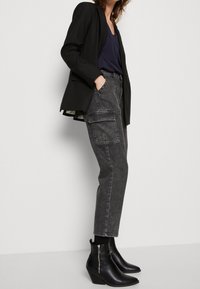 Graue Cargo-Hosen mit seitlichen Taschen, getragen mit einem schwarzen Blazer und figurbetonten schwarzen Ankle-Boots. Das Outfit besteht aus einem navy-blauen Oberteil darunter.