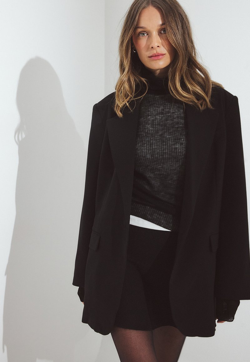 Blazer negro de gran tamaño hecho de una tela suave con solapas muescas, llevado sobre una parte superior texturizada oscura y una falda negra ajustada.