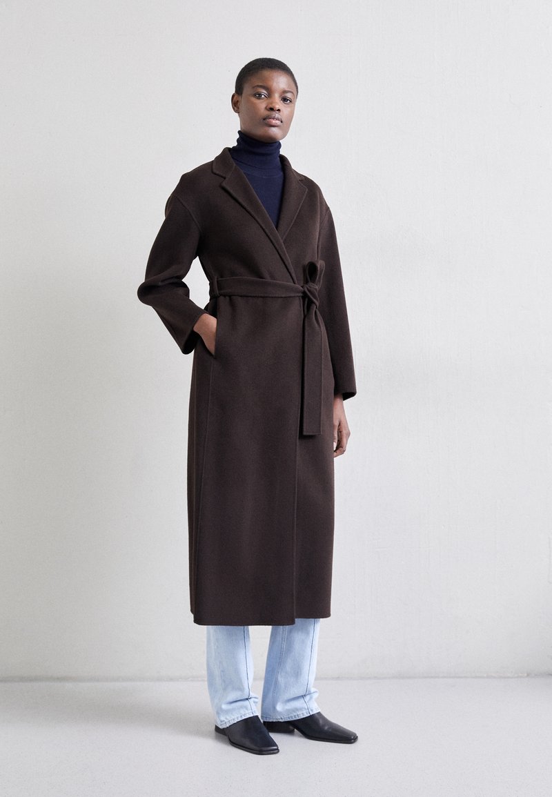 Brun indpakningsfrakke med en bælte, lange ærmer og sjalskrave, parret med en sort turtleneck og lyseblå jeans.