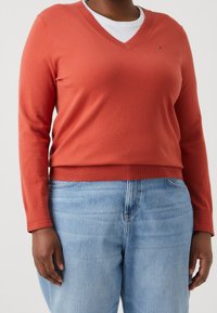 Pull à col en V en mélange de coton terracotta, avec manches longues et poignets côtelés, porté avec un jean bleu clair. Petit logo sur la poitrine.