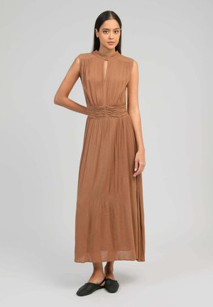 Femme debout, un bras plié, portant une robe maxi marron sans manches et des chaussures plates tissées noires, devant un fond gris uni.