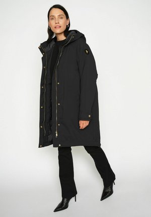 Schwarze lange Parkajacke mit Kapuze, goldenen Details und vorne mit Reißverschluss. Hat eine glatte Textur und seitliche Taschen. Wird über schwarzen Hosen getragen.