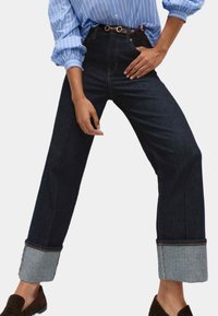 Persona che indossa jeans blu scuro con risvolti, una cintura marrone, una camicia a righe blu con le maniche arrotolate e scarpe scamosciate marroni scure.