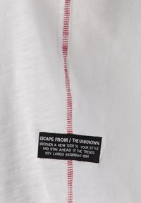 Tessuto bianco con cuciture verticali rosse e un'etichetta nera con la scritta "Fuggi dall'ignoto, scopri un nuovo lato del tuo stile, Key Largo 1994."