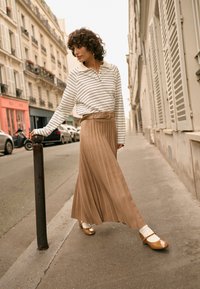 Camicia a maniche lunghe a righe abbinata a una gonna marrone plissettata con cintura. Scarpe in vernice color cuoio e calzini bianchi. Ambientazione in una strada urbana.