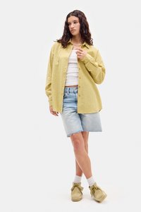 Żółta koszula w kratę, biały top na ramiączkach, jasnoniebieskie szorty dżinsowe, beżowe buty, postawa stojąca, jednolite tło, luźny krój, warstwowy wygląd.