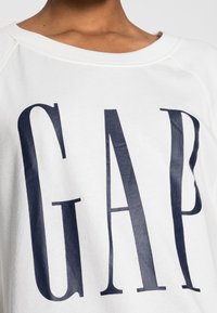 Felpa bianca con un grande logo "GAP" blu navy sulla parte anteriore, dal fit rilassato e con scollatura rotonda. Tessuto in cotone con una texture liscia.