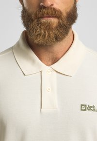 Crème polo shirt met een textuurpatroon, twee knopen en een groene Jack Wolfskin-logo aan de linkerkant van de borst.