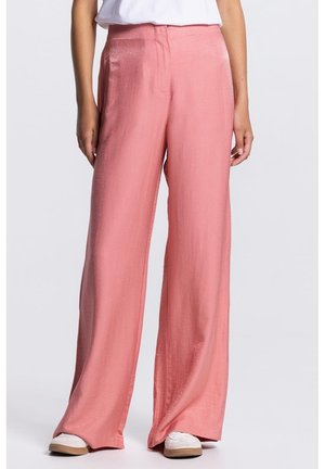 Vrouw die een loszittende satijnen roze broek met hoge taille en brede pijpen draagt, gecombineerd met witte sneakers en een wit, ingestopt shirt.