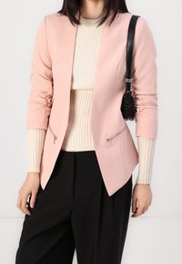 Světle růžový blazer s přiléhavým střihem, obsahující kapsy na zip a dlouhé rukávy, položený na krémovém žebrovaném svetru a černých kalhotách.