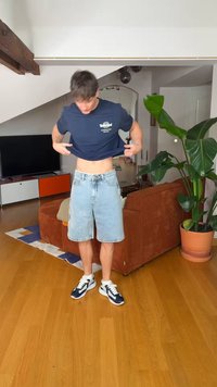 Jeune homme en chemise bleue et short en jean soulevant sa chemise dans un salon avec un sol en bois, une grande plante, une télévision et un canapé marron.