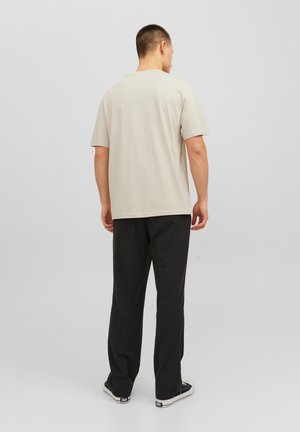 Beige T-Shirt mit kurzen Ärmeln und geradem Schnitt, kombiniert mit schwarzen Hosen und Stoffschuhen, von hinten betrachtet. Schlichte, einfarbige Gestaltung.