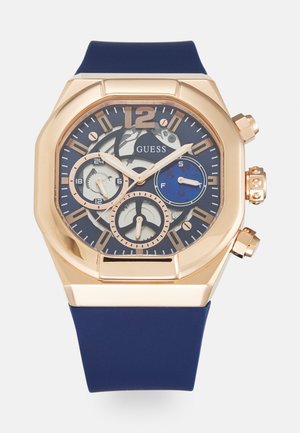 Goldfarbene, achtkantige Uhr mit navyfarbenem Silikonarmband, blau-weißem Ziffernblatt, mehreren Teilzifferblättern und silbernen Akzenten auf den Stundenmarkierungen und Zeigern.