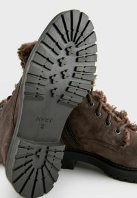 Botas de tobillo de ante marrón con un borde de pelo texturizado, que cuentan con una suela de goma negra resistente con ranuras para tracción y ojales reforzados para los cordones.