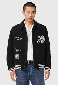Mand i sort varsity jakke med hvide lapper, stribede manchetter, hvid skjorte, blå jeans og sort bælte, som står foran en ensfarvet baggrund.