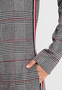 Tissu écossais gris et noir avec des accents rouges, comportant une poche latérale et des manches longues avec un design à poignet. Matériau en tricot texturé.