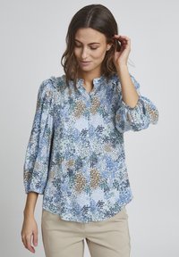 Svetlomodrá blúzka s kvetinovým vzorom v zelených, hnedých a modrých tónoch. Má zapínanie na gombíky vpredu a vypchaté rukávy s volánovými okrajmi.