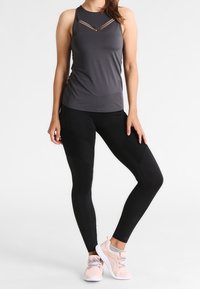 Grijs sporttop met uitsnijdingen op de borst, gecombineerd met zwarte legging met textuurelementen. Lichtroze sportschoenen maken de uitstraling af.