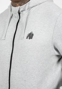 Grijze zip-up hoodie van zachte stof, met een zwarte rits en een klein zwart gorilla-logo op de linkerkant van de borst.