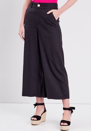 Frau trägt hoch taillierte, weite schwarze Caprihosen mit Fronttaschen und schwarzen Plateau-Sandalen mit Knöchelriemen.