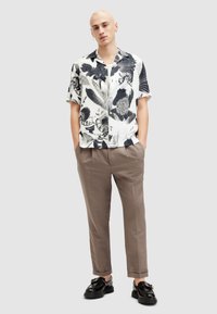 Hombre con una camisa de manga corta de patrón abstracto en blanco y negro, pantalones taupe y mocasines negros, de pie con las manos en los bolsillos.