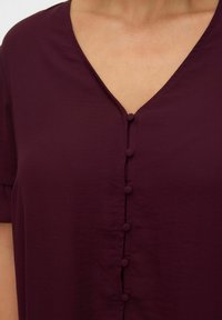 Burgundská blůza s výstřihem do V, krátkými rukávy a řadou kulatých knoflíků na přední straně pro zapínání. Hladká textura látky.