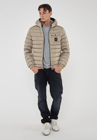 Giubbotto imbottito beige con cappuccio, zip frontale e tasca sul petto. Abbinato a t-shirt grigia, jeans scuri e sneakers bianche. Texture liscia.