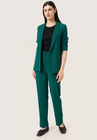 Tyrkysový blazer s ohrnutými rukávy a odpovídajícími kalhotami na míru, v kombinaci s černým upnutým topem. Jemná textura, elegantní linie a minimální akcenty.