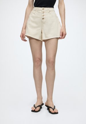 Person trägt hochgeschnittene beige Shorts mit Knöpfen vorne, ein schwarzes ärmelloses Top und schwarze Sandalen mit Absatz, steht vor einfarbigem Hintergrund.