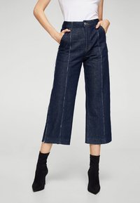 Pantalones recortados de mezclilla oscura con costuras visibles, dos bolsillos laterales, una cintura alta y un corte recto, combinados con botas negras hasta el tobillo.