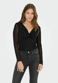 Giovane donna con capelli lunghi che indossa un top nero trasparente a maniche lunghe con riflessi e jeans scuri a vita alta, posando davanti a uno sfondo bianco.