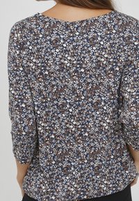 Blouse à imprimé floral avec un fond bleu marine, ornée de fleurs blanches, bleues et brunes. Le design comprend des manches trois-quarts et un décolleté en forme de scoop.