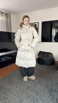 Dunjakke i lys kremfarge med belte i livet, hette med pelsfôr og to frontlommer. Myk tekstur, knelengde, kombinert med mørke bukser og støvler.