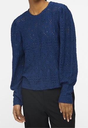 Femme portant un chemisier en dentelle bleu marine à manches longues bouffantes et un pantalon noir sur fond blanc.