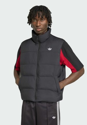 Mand med korte dreadlocks iført en sort polstret vest over en rød og sort kortærmet skjorte og sorte Adidas træningsbukser med hvide striber.
