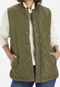 Mujer con un chaleco acolchado verde oliva y botones a presión sobre un suéter de punto blanco y jeans azules, con las manos en los bolsillos del chaleco.