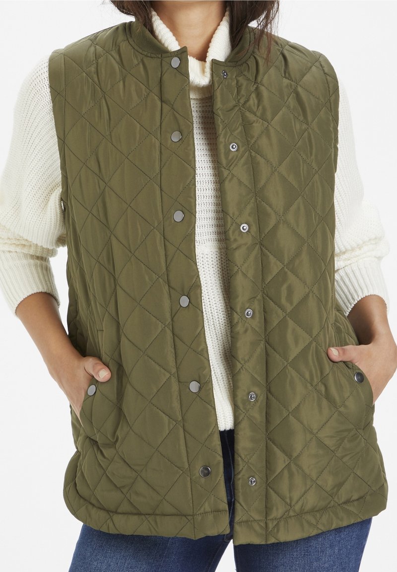 Mujer con un chaleco acolchado verde oliva y botones a presión sobre un suéter de punto blanco y jeans azules, con las manos en los bolsillos del chaleco.