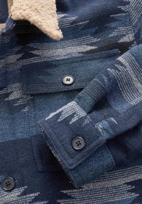 Kabát z vlněné směsi, který má tmavě modrý a béžový vzor, s měkkým fleecovým límcem a manžetami na knoflíky. Pozoruhodný kontrast textur.