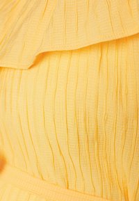 Vestido amarelo com um tecido texturizado, apresentando pregas verticais e um decote frouxo. Tem uma cintura definida acentuada por um cinto coordenado.
