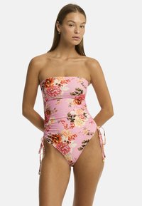 Un mannequin pose dans un maillot de bain floral rose avec un design sans bretelles et des nœuds sur les côtés, affichant une expression naturelle et confiante.