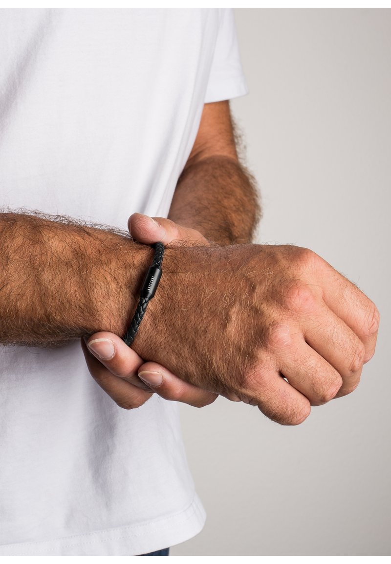 Les bras et mains poilus d’un homme ajustant un bracelet en cuir tressé noir, portant une chemise blanche à manches courtes sur un fond uni.