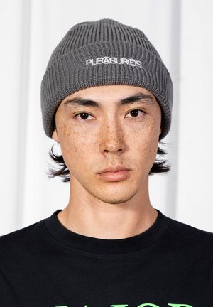 Młoda osoba z jasnymi piegami, nosząca ciemnoszary beanie z napisem "PLEASURES" oraz czarną bluzę bez kaptura na jednolitym białym tle.