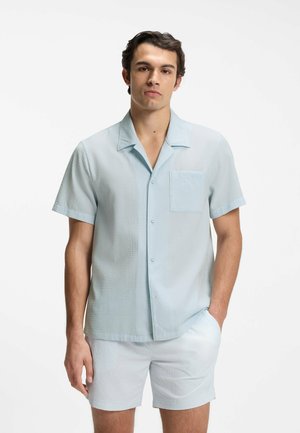 Mann trägt hellblaues, strukturiertes Kurzarmhemd mit Knöpfen und passende Shorts, steht mit einer Hand in der Tasche vor weißem Hintergrund.
