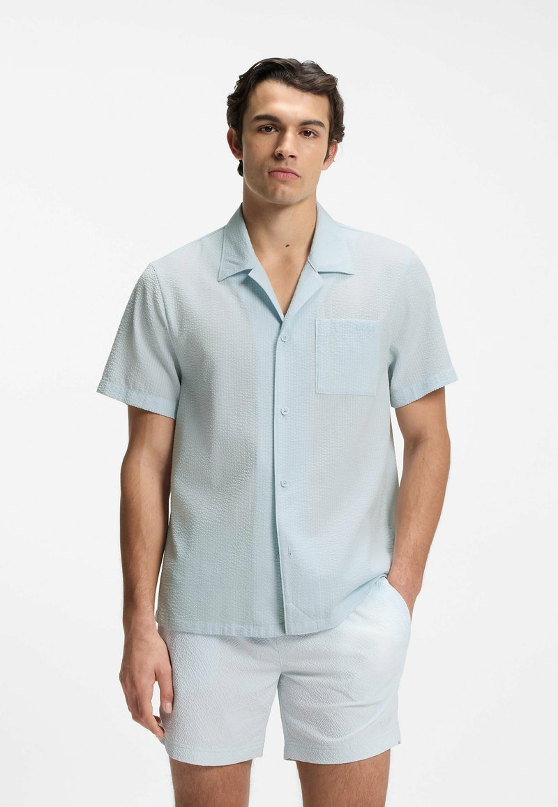 Homme portant une chemise texturée bleu clair à manches courtes boutonnée et un short assorti, debout avec une main dans la poche sur fond blanc.