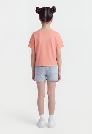 Child with two braided buns wearing a peach short-sleeve shirt, light denim shorts, white socks, and white sneakers, standing facing away.