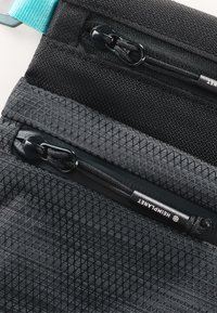 Tessuto nero testurizzato con motivo a diamante; presenta due tasche con zip e una tracolla turchese per il fissaggio. Etichetta del marchio visibile sulla superficie.