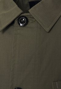 Olivgrön plagg med en strukturerad krage och en blank svart knapp. Har en texturerad tyg med subtila linjer och kontrasterande sömmar.