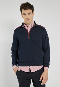 Granatowy sweter z pół-zamkiem i czerwonymi akcentami, noszony na różowej koszuli w paski. Posiada ściągacze przy mankietach i u dołu, a także kołnierz.